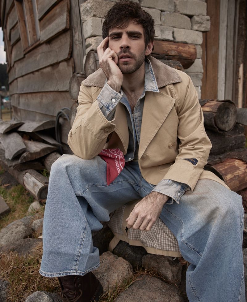 Portrait of a man with dark hair, wearing a tan coat and wide-leg jeans, sitting outdoors with a building in the background.