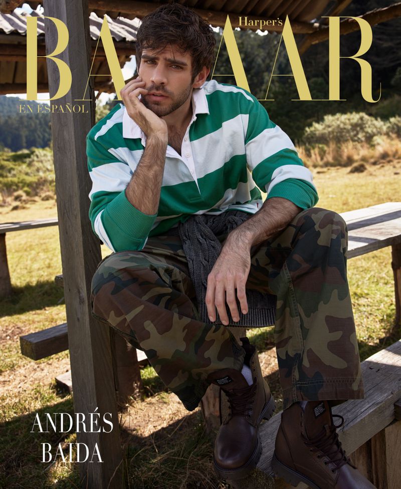 Man in a Harper's Bazaar fashion editorial, wearing a rugby shirt and camouflage pants, poses outdoors with mountain views.