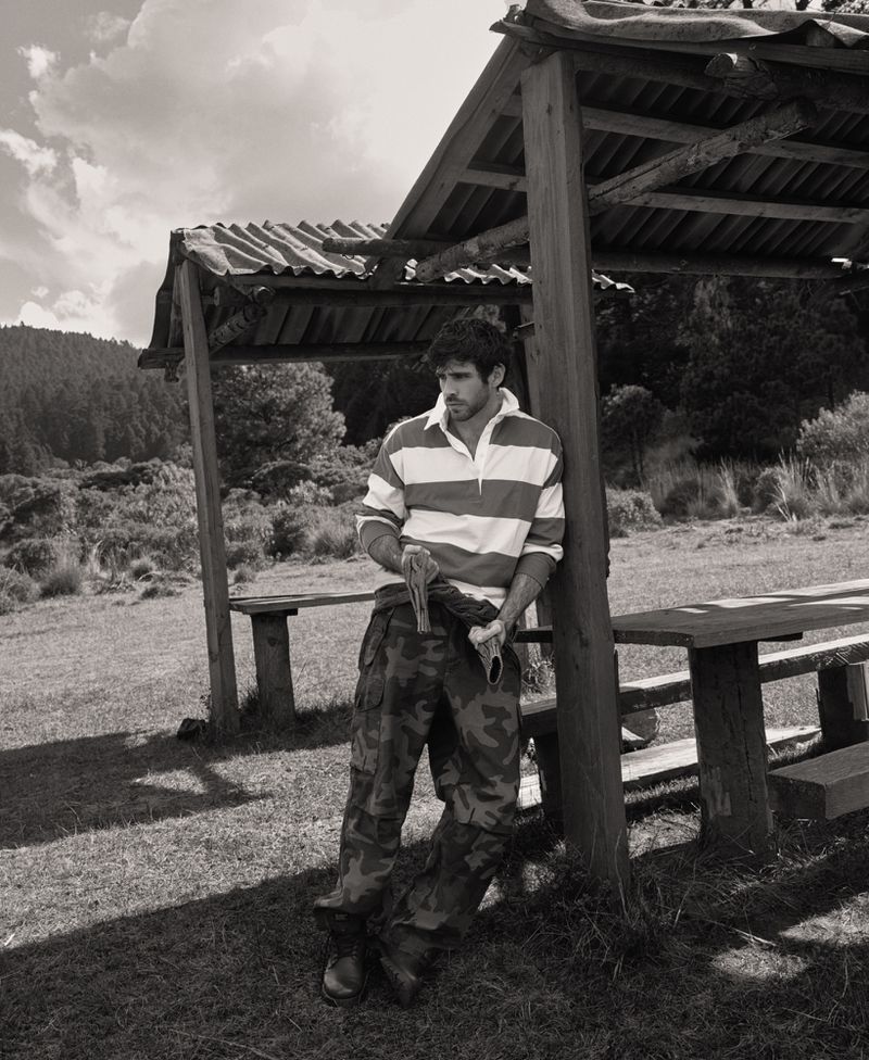 Monochrome outdoor shot of a man in a striped shirt and camouflage pants leaning against a wooden structure with a forest background.