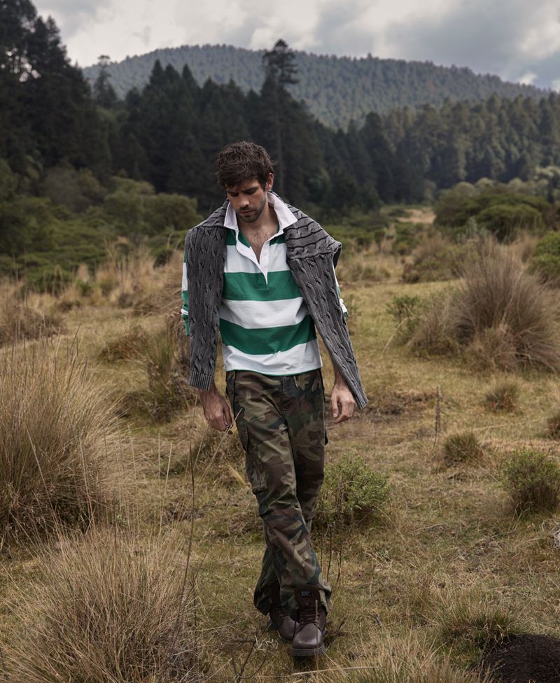 Male fashion model in outdoor shot wearing casual attire in a field with mountains and trees in background. Natural light, full shot.