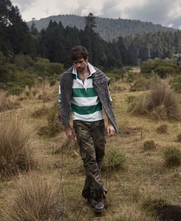 Male fashion model in outdoor shot wearing casual attire in a field with mountains and trees in background. Natural light, full shot.