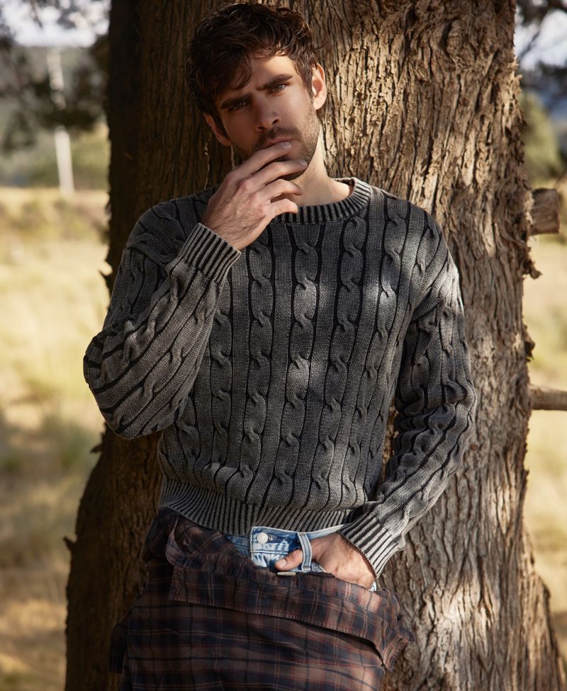 Portrait of a bearded man in a cable-knit sweater leaning against a tree trunk in an outdoor setting with soft natural light.