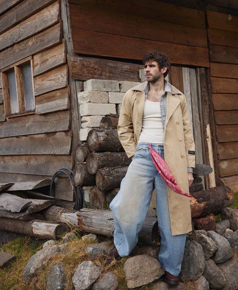 A male model poses outdoors in a tan trench coat and light wash jeans against a rustic wooden building and a pile of logs.