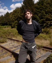 Man standing on railroad tracks wearing a black jacket and grey jeans against a green forest background