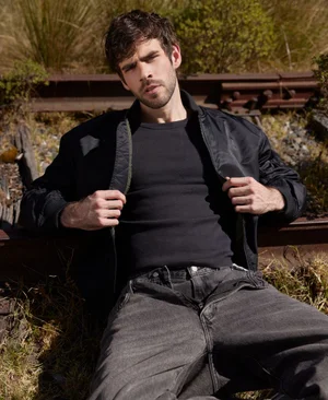 Man with dark hair wearing a black jacket and grey jeans seated near railway tracks outdoors.