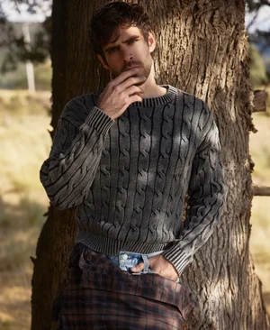 Man wearing a grey cable-knit sweater standing outdoors against a tree trunk for Harper's Bazaar Mexico editorial.