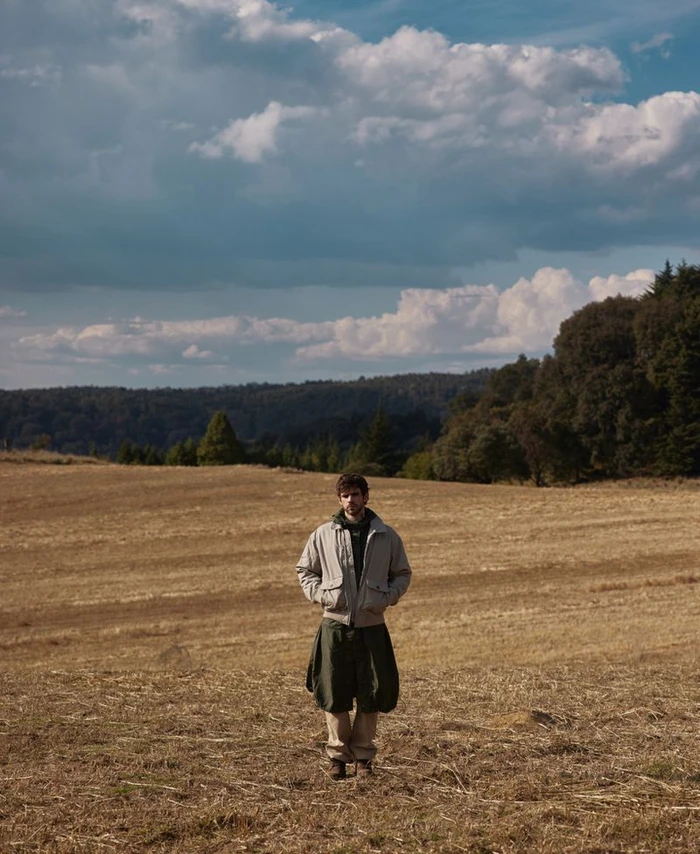 A man wearing a grey bomber jacket and dark trousers standing in a wide dry field under a dramatic cloudy sky.