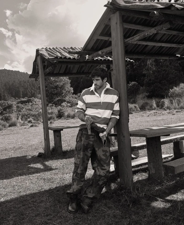 Man wearing a striped rugby shirt and camo pants leaning against a wooden shelter outdoors