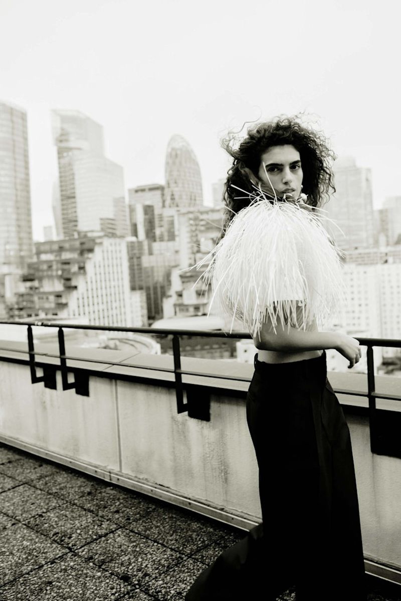Fashionable adult with curly hair wearing a feathered boa walks on a rooftop with a city skyline in the background in a black-and-white photograph.