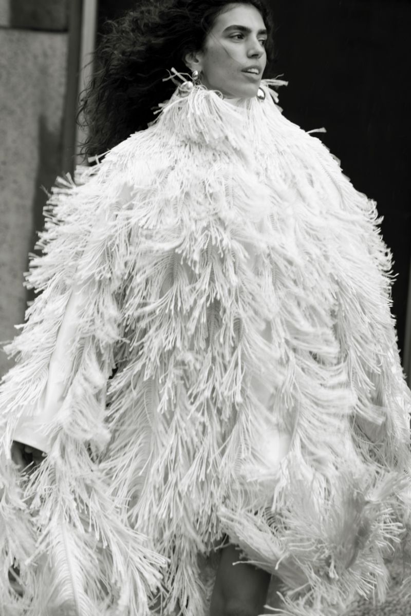 Model wearing a textured white fringed coat against a textured architectural background in a black and white fashion photograph.
