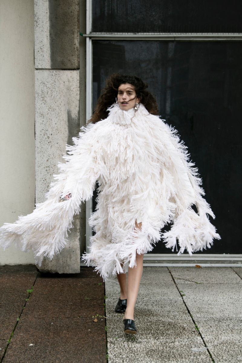Model wearing a textured white fringed turtleneck garment walking outdoors on stone pavement.