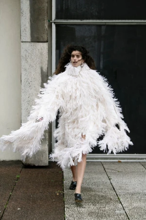 Model wearing a textured white fringed turtleneck garment walking outdoors on stone pavement.