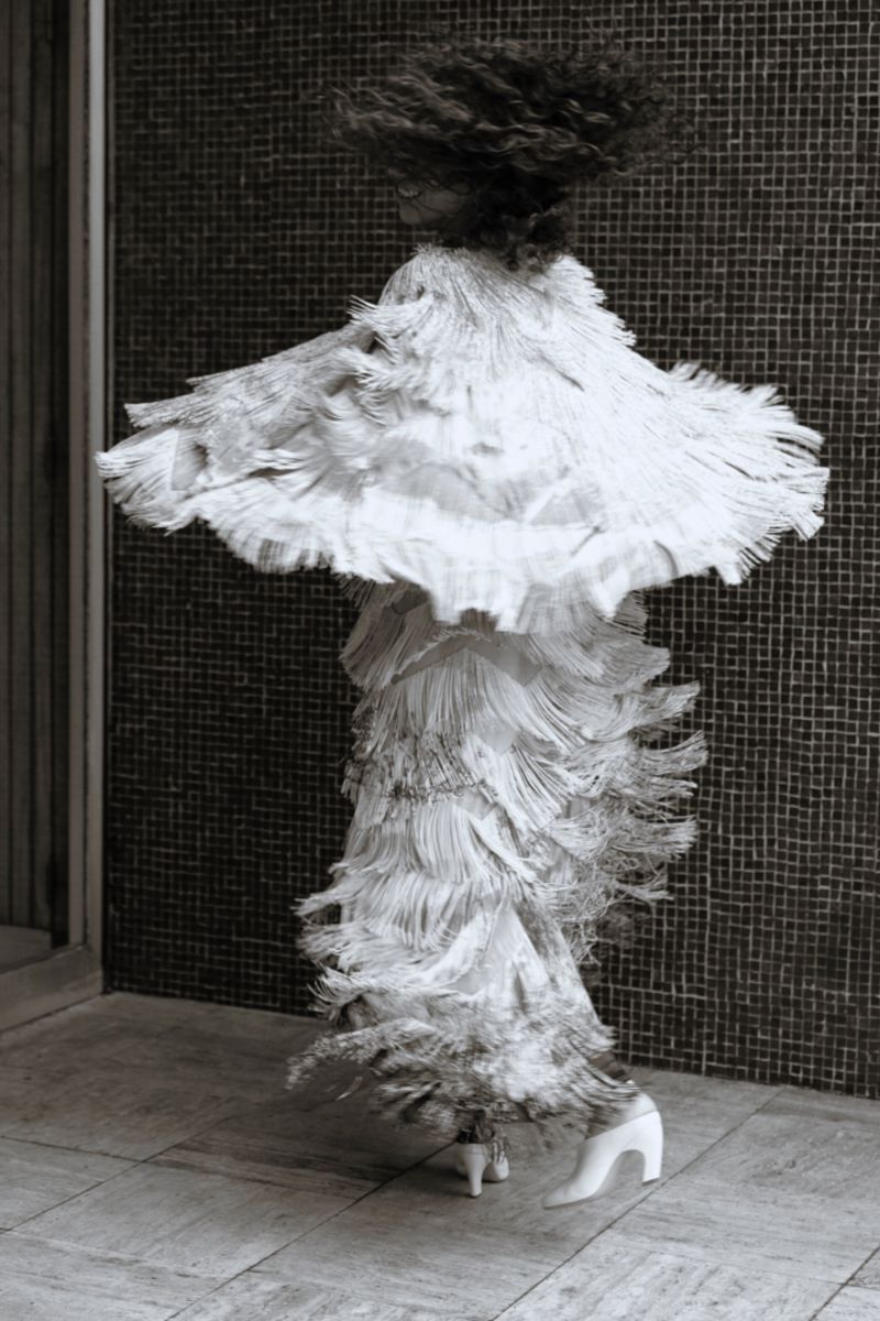 Monochrome fashion photography: Adult female model in feathered dress and high heels dances in motion on a tile floor near a stone wall.