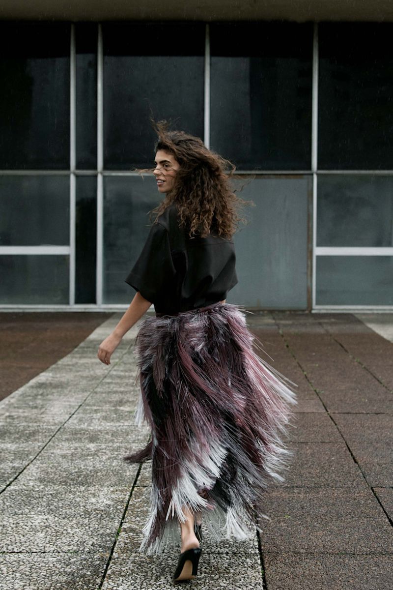 Model with long curly hair wearing a black top and textured fringe skirt walking outdoors