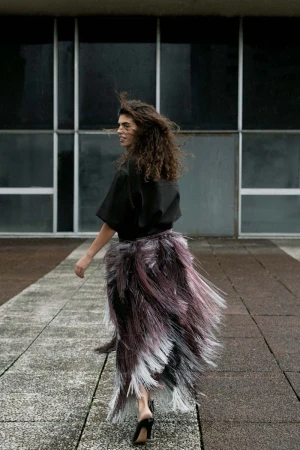 Model with long curly hair wearing a black top and textured fringe skirt walking outdoors
