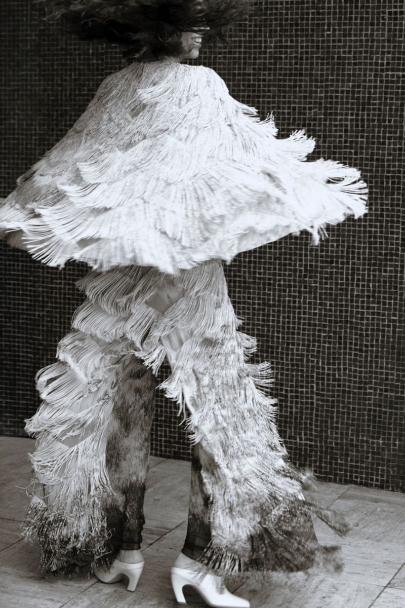 Monochrome shot of person in elaborate fringed outfit in motion, captured against a textured wall. Elegant shoes complete the look.