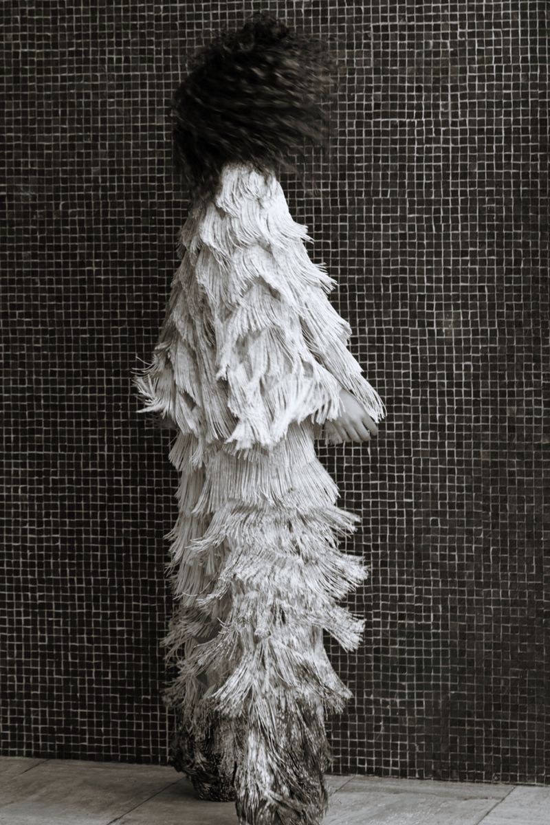 Monochrome artistic studio shot of a person in an elaborate fringed outfit, standing against a textured tile background, creating a unique silhouette.