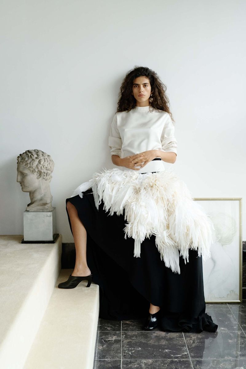 Elegant woman in fashionable outfit poses against a white wall next to a classical bust, showcasing beauty and high-end clothing.
