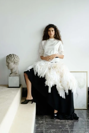 Elegant woman in fashionable outfit poses against a white wall next to a classical bust, showcasing beauty and high-end clothing.