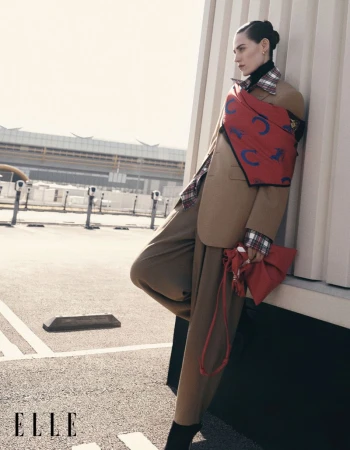 Fashion editorial shot: Adult female model poses in oversized brown suit, red purse, and plaid top near urban roadside with building.
