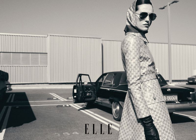 Fashion shot of a woman in a patterned coat, headscarf, and sunglasses, standing next to a vintage car on an outdoor lot in black and white.