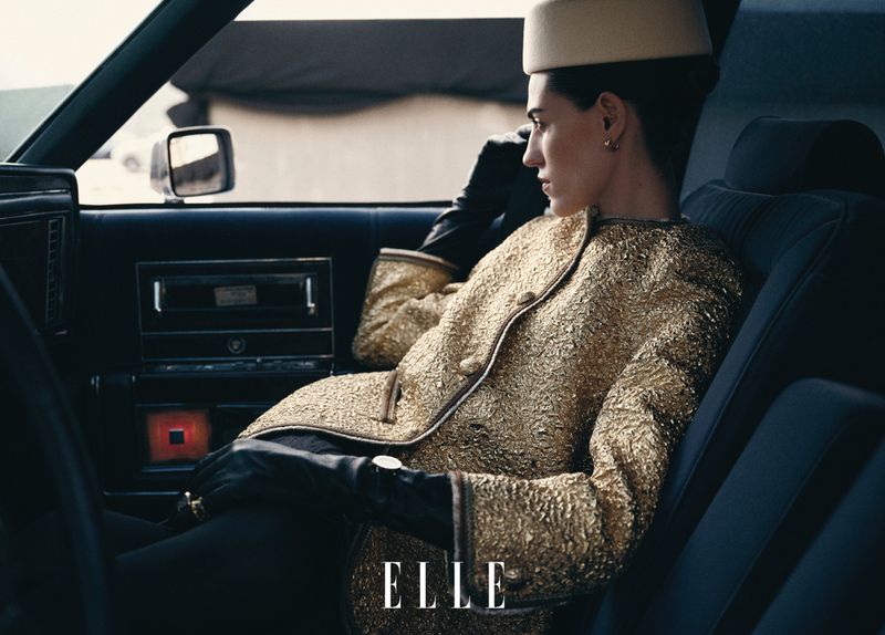 Stylish woman in gold textured jacket and cream hat sits in a car interior with black leather, giving off a fashionable, retro aesthetic.