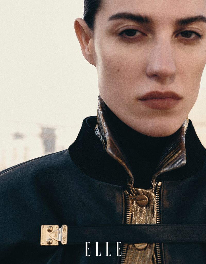 Close-up of a woman in a black jacket and dark turtleneck, posing in a neutral outdoor setting with the ELLE magazine logo visible.