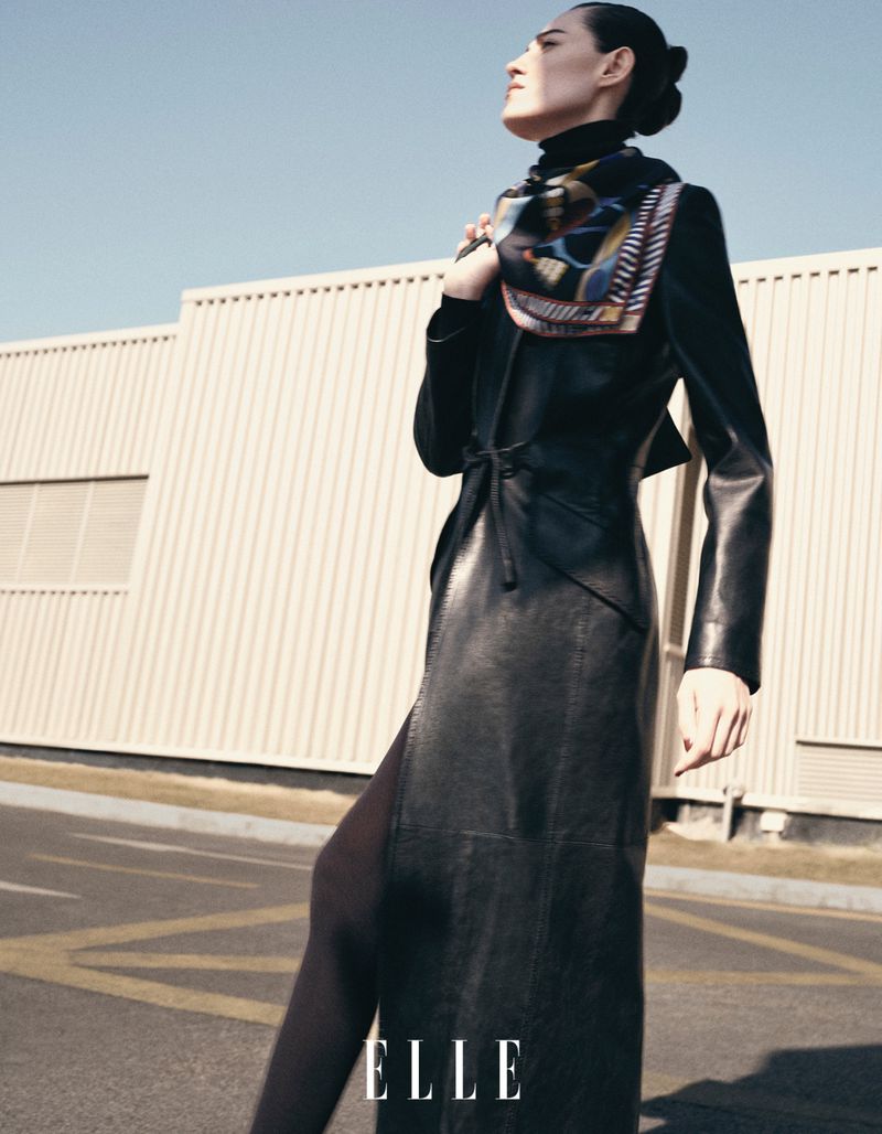 Woman in long black coat and patterned scarf walks on city street with industrial building backdrop. Fashion editorial style.