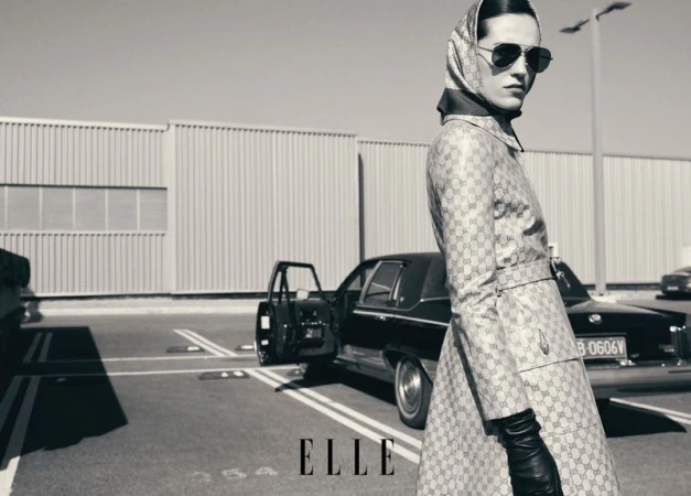 Model in patterned trench coat and headscarf with sunglasses posing outdoors near a classic car in an ELLE China editorial.