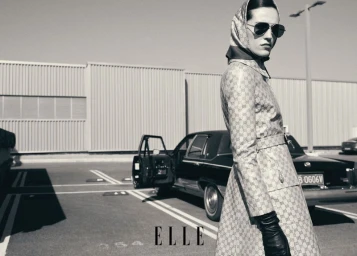 Model in patterned trench coat and headscarf with sunglasses posing outdoors near a classic car in an ELLE China editorial.