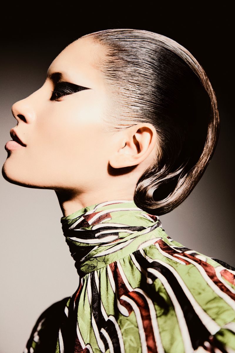 Glamour shot of a stylish woman with slicked-back hair and bold eye makeup, wearing a patterned green, white, and brown top.