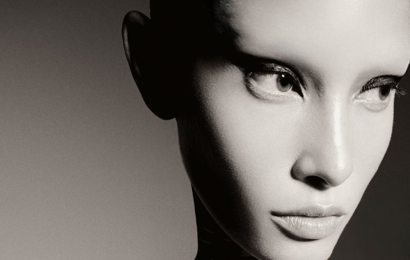 Close up monochrome portrait of a model with dramatic winged eyeliner against a plain dark grey studio background.