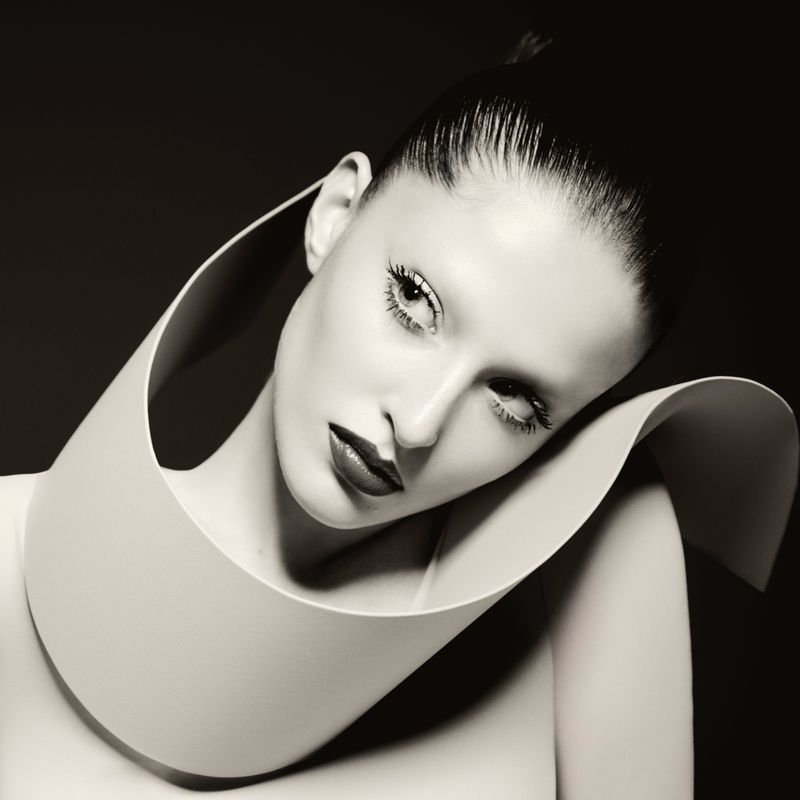 Striking monochrome portrait of a woman with slicked-back hair, bold makeup, and a large, sculptural white collar against a dark backdrop.