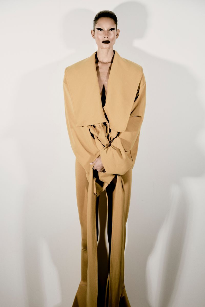 Adult female fashion model posing in a full-length gold garment in front of a plain white wall, artistic makeup and slicked-back hair.