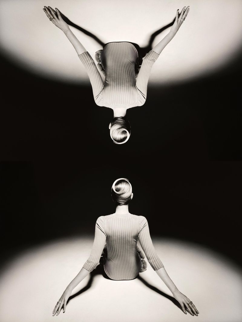 Mirrored studio shot of an adult woman with her arms raised above her head. She wears a ribbed top. The background is gradated.