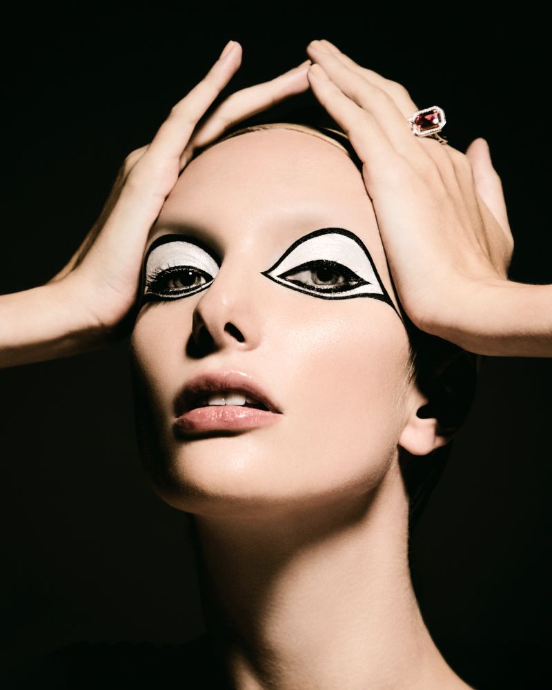 Glamour shot of a woman with white and black makeup design around her eyes, wearing a ring, set against a dark background.