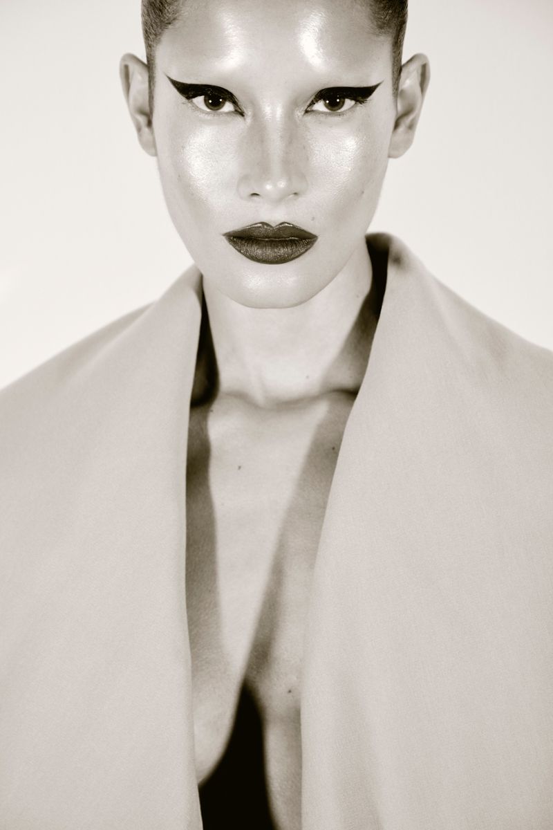 Close-up portrait of a young woman with dramatic black winged eyeliner and dark lipstick against a white background in a studio setting.