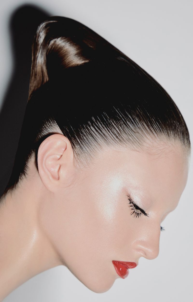 Close-up studio portrait of a woman with slicked-back hair, red lipstick, and minimal eye makeup against a bright white background.