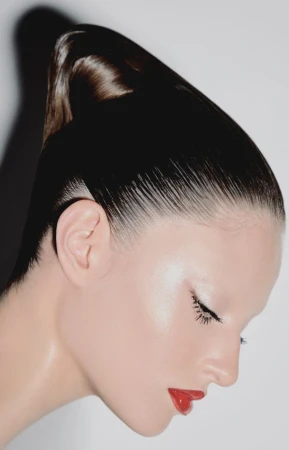 Close-up studio portrait of a woman with slicked-back hair, red lipstick, and minimal eye makeup against a bright white background.