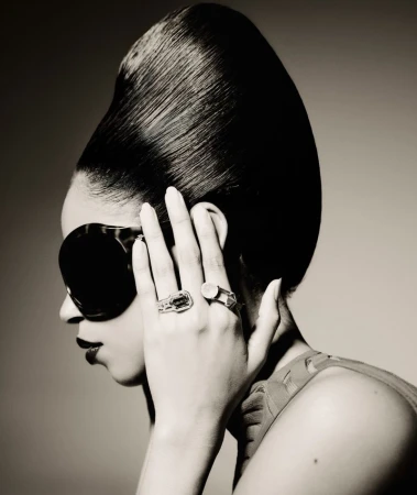 Portrait of an adult woman with an elegant hairstyle and sunglasses, wearing rings, in a studio with soft, neutral lighting.