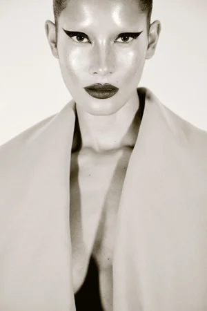 Close-up portrait of a model with sharp winged eyeliner and dark lips wearing a structured coat against a plain background.