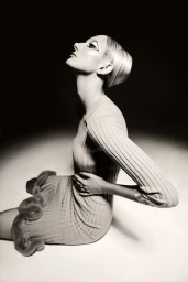 Monochrome portrait of a woman with graphic eye makeup seated against a white studio background wearing a ribbed knit dress