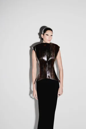 Model with dark winged eyeliner and sleek dark hair wearing a structured metallic top and black skirt against white seamless background.