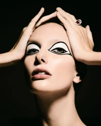 Close-up portrait of a woman with bold graphic eye makeup holding her hands to her head against a dark background