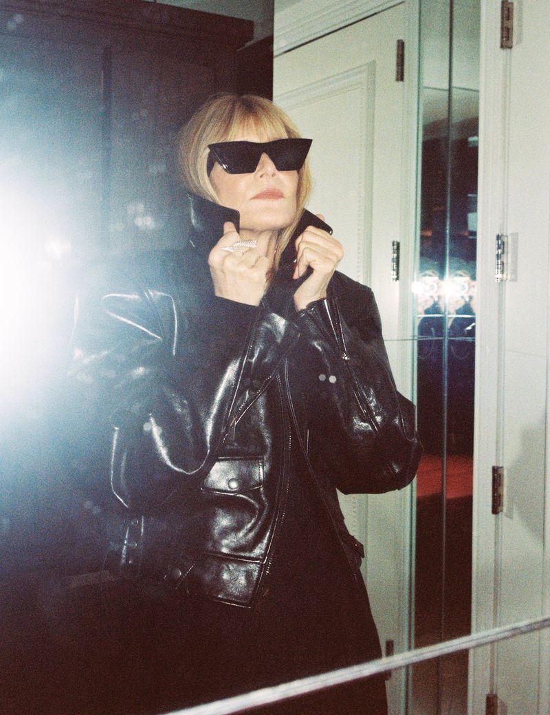 Elegant woman with black sunglasses and leather jacket in an indoor setting, posing confidently with a slight smile.