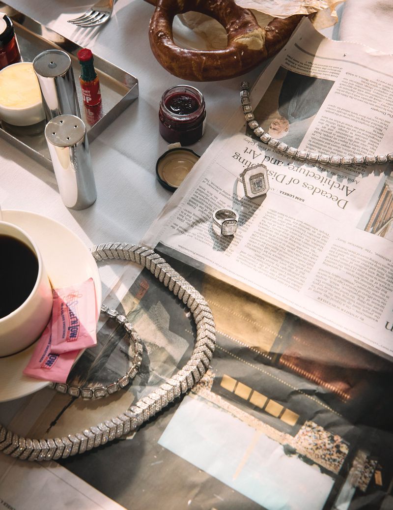 High-angle shot of an editorial fashion still life featuring jewelry, food, coffee, and a newspaper on a white surface.