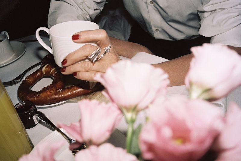 Person holding a mug with a large ring, pretzel, jam, and flowers on a table. Focus on food, hand jewelry and tabletop setting.