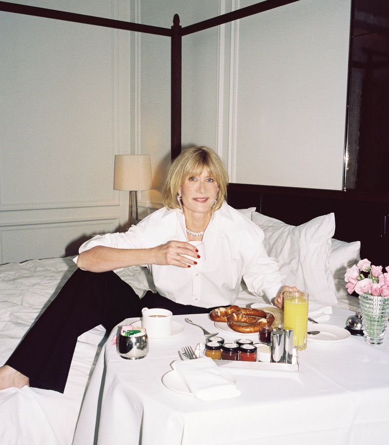 Woman in white shirt and black pants sits on bed with a breakfast tray featuring pastries, coffee, and juice in a well-lit room.