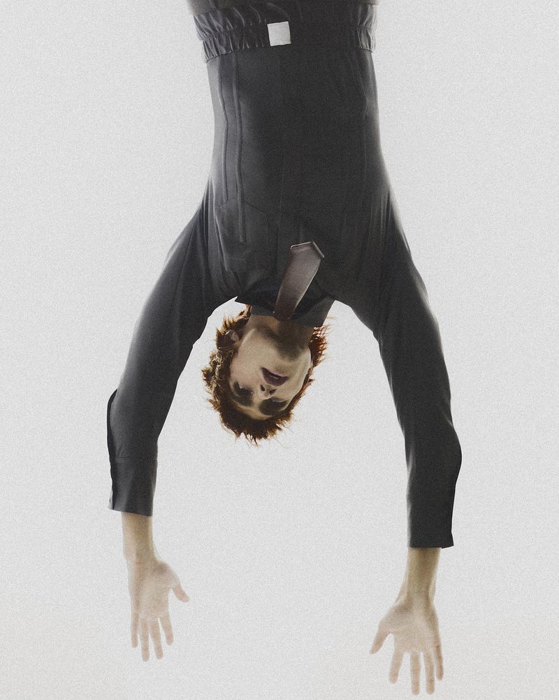 Upside-down portrait of an adult male wearing a dark suit and tie against a stark white background in a dramatic studio lighting setup.
