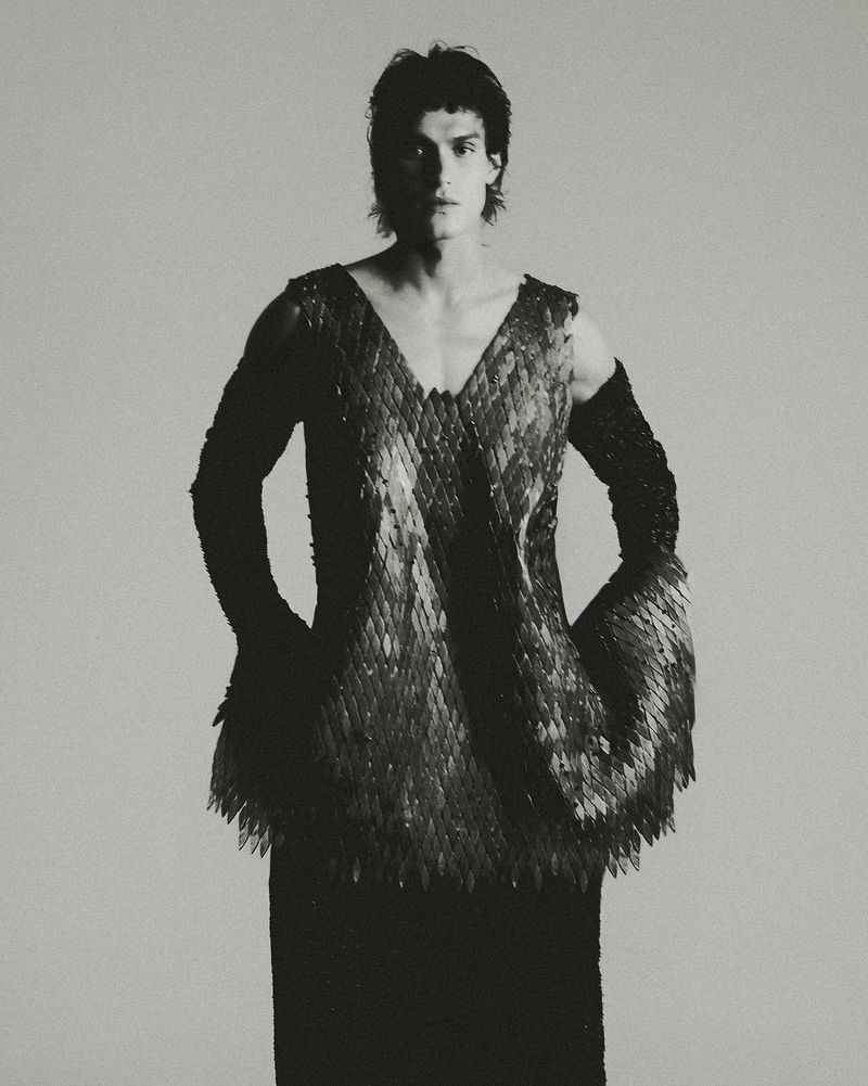 Monochrome portrait of a male model wearing a unique v-neck top with scale-like embellishments and fitted black arm sleeves, against a light grey backdrop.